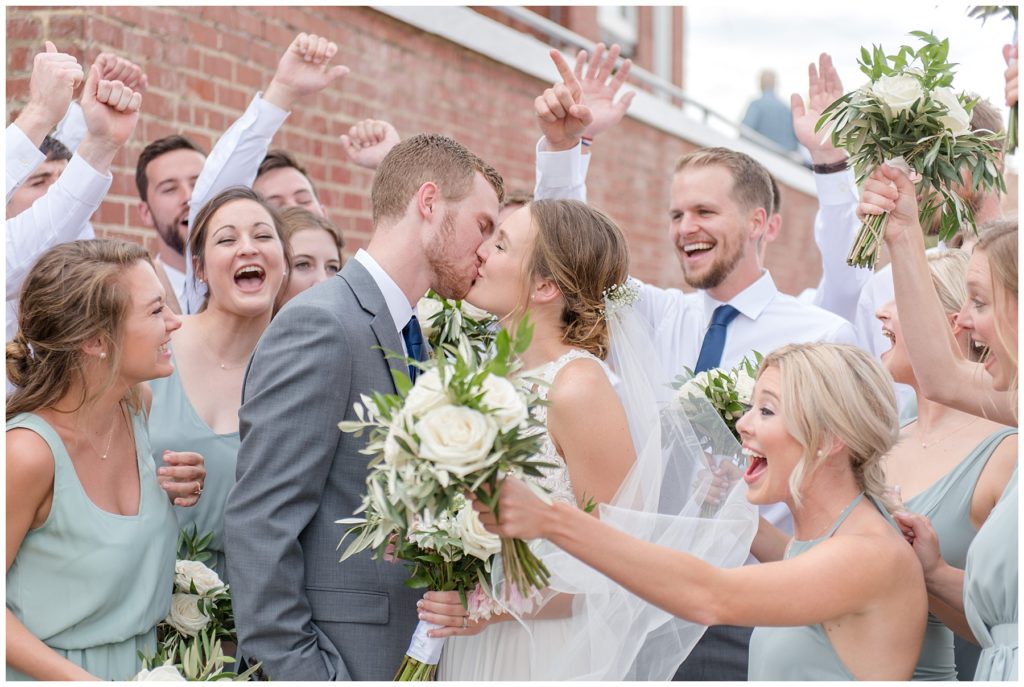 Bride and groom kissing while surrounded by their cheering bridal party on their wedding day